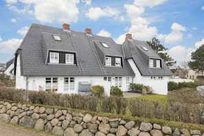 A two-story house with a steeply pitched roof, dormer windows, and a stone wall in the foreground. - Friesenglück - 3-room vacation apartment for up to 4 people with sunbathing area. (Sylt)