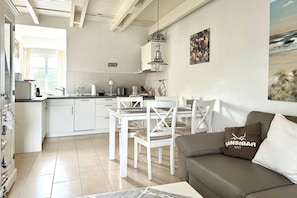 A modern kitchen with white cabinets, a dining area with white chairs, and a grey sofa with a cushion. - Friesenglück - 3-room vacation apartment for up to 4 people with sunbathing area. (Sylt)