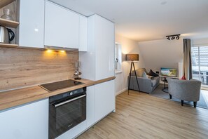 Een moderne keuken met witte kasten en een houten aanrecht, een inbouwoven, en een zithoek met een grijze bank en een vloerlamp.