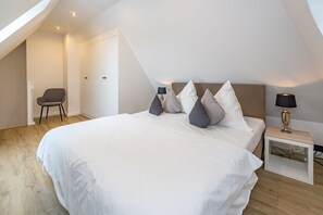 Een slaapkamer met een groot bed, een stoel, een nachtkastje met een lamp en een schuine kap.