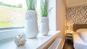 Ein Schlafzimmer mit einem Bett, einem Nachttisch, einer Lampe und zwei weißen Vasen mit Pflanzen auf einem Fensterbrett.