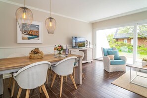 Un comedor con mesa de madera, sillas de mimbre, televisor y vistas al jardín a través de amplias ventanas.