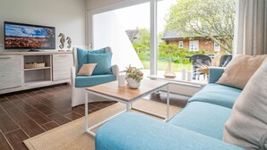 Ein modernes Wohnzimmer mit Sofa, Sessel und Couchtisch. Ein Fernseher steht auf einem Lowboard, ein Kamin ist vorhanden und ein großes Fenster bietet Blick auf Garten und Haus.