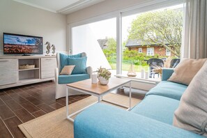 Ein modernes Wohnzimmer mit Sofa, Sessel und Couchtisch. Ein Fernseher steht auf einem Lowboard, ein Kamin ist vorhanden und ein großes Fenster bietet Blick auf Garten und Haus.