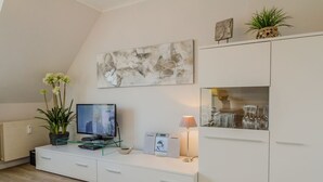 Un salon moderne avec un téléviseur à écran plat, une table basse en verre, un meuble blanc et une plante décorative.