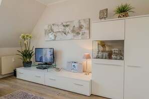 Un salon moderne avec un téléviseur à écran plat, une table basse en verre, un meuble blanc et une plante décorative.