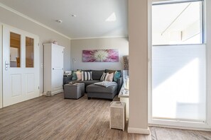 A spacious living area with a grey sofa, ottoman, and a large window providing natural light. - Schloss am Meer, App. 5 "Strandläufer" - 3-room vacation apartment with terrace - (Wenningstedt-Braderup (Sylt))