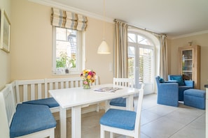 A dining area with a white table and blue upholstered chairs, a vase of flowers, and a view of the outside through large windows.