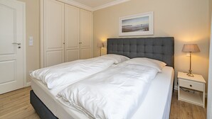 En pænt redt seng med hvide sengetæpper, en grå polstret sengegavl og et natbord med en lampe.