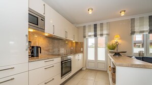 Una cocina moderna con armarios blancos, horno empotrado, microondas y cafetera. Hay una ventana con persianas y una planta en la encimera.