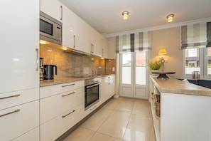 A modern kitchen with white cabinetry, a built-in oven, a microwave, and a coffee maker. There is a window with blinds and a plant on the counter.
