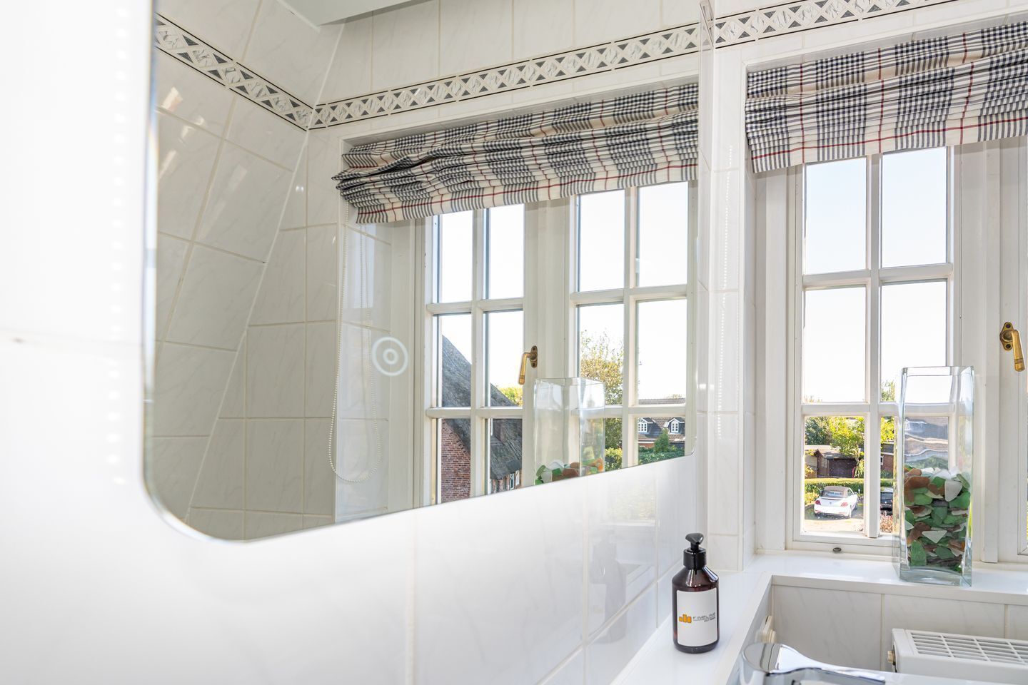 A bathroom with white tiles, a large mirror, and a window with a checkered blind.