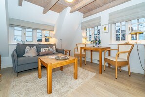 A cozy living room with a sofa, coffee table, and dining table, featuring wooden beams and large windows. - Altstadttor App. 14 - Central 2-room apartment under thatch in Alt (Sylt)