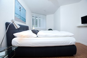 Ein modernes Schlafzimmer mit Bett, Schreibtisch mit Lampe, Wand-TV und Fenster mit Jalousien.