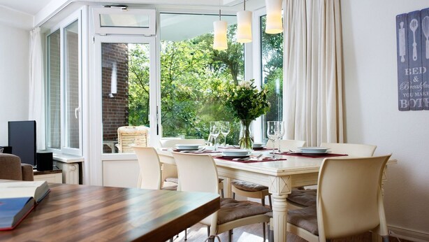 Un comedor con una mesa dispuesta para cuatro personas, un televisor sobre un mueble bajo y una ventana con cortinas.