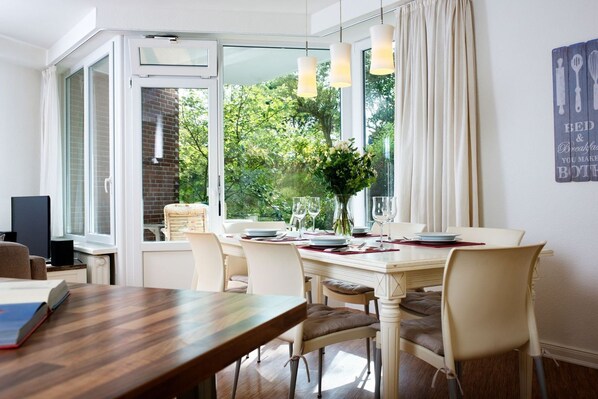 Un comedor con una mesa dispuesta para cuatro personas, un televisor sobre un mueble bajo y una ventana con cortinas.