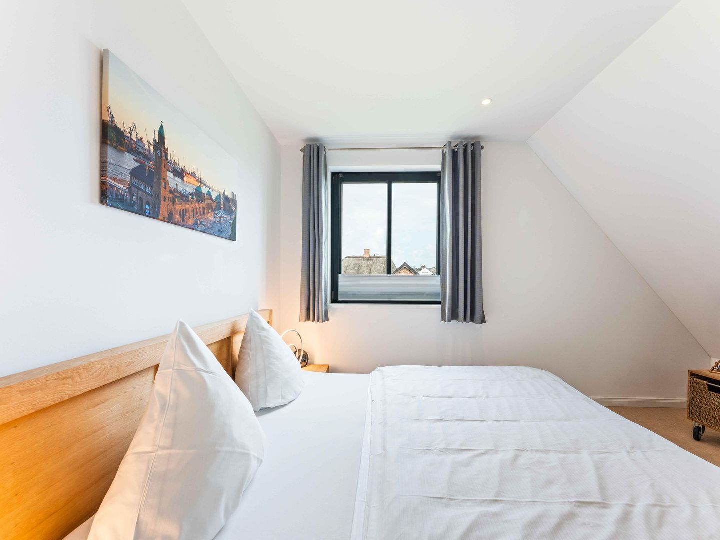 Un dormitorio con cama, mesita de noche, ventana con cortinas y un cuadro de un paisaje urbano en la pared.