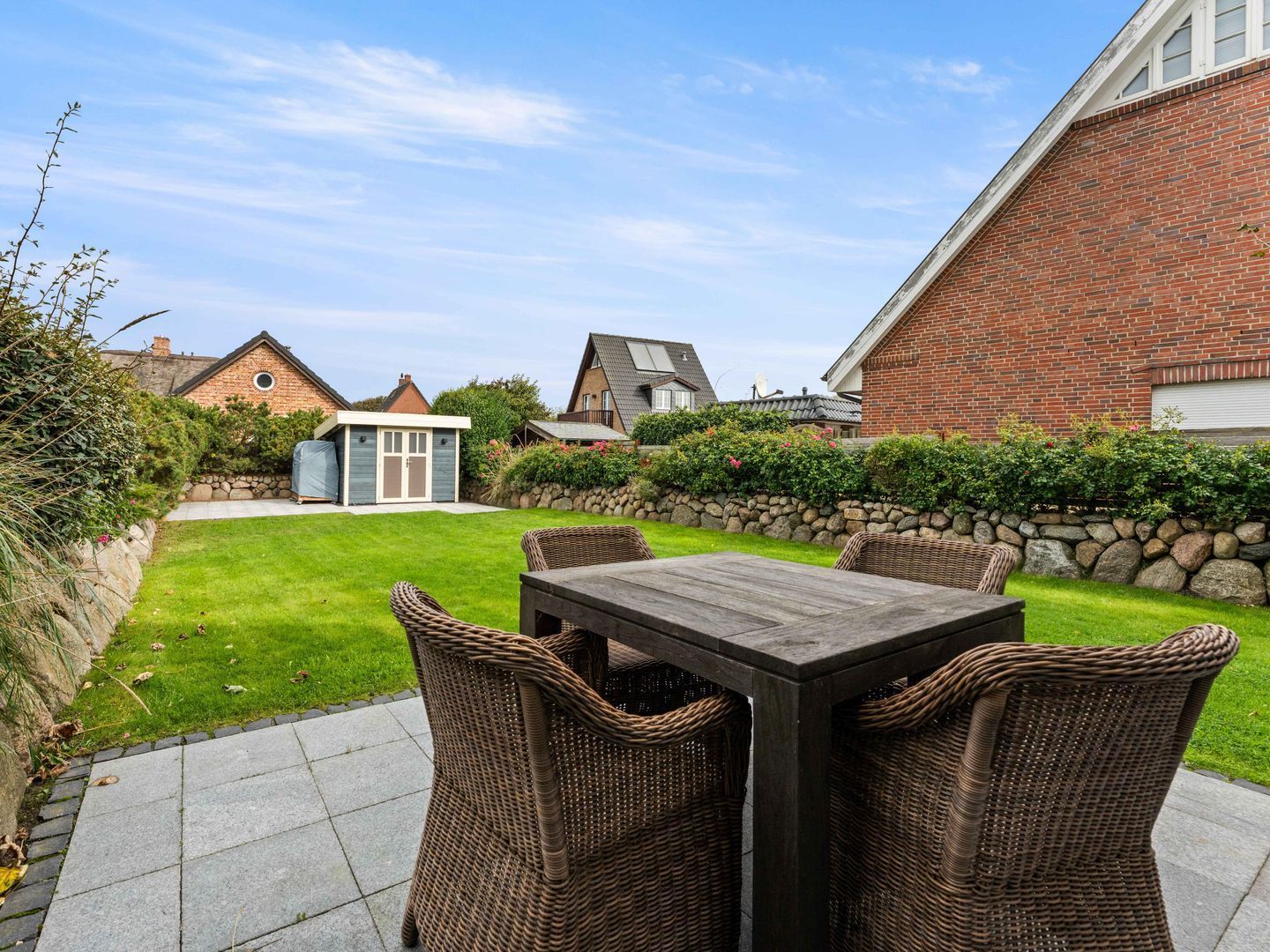 A patio with a wooden table and wicker chairs, surrounded by a well-maintained lawn and a stone wall.