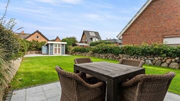 A patio with a wooden table and wicker chairs, surrounded by a well-maintained lawn and a stone wall.