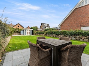 A patio with a wooden table and wicker chairs, surrounded by a well-maintained lawn and a stone wall.