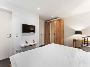 Habitación de hotel con una cama, televisor montado en la pared, armario de madera, mesita de noche con lámpara y escritorio con computadora.