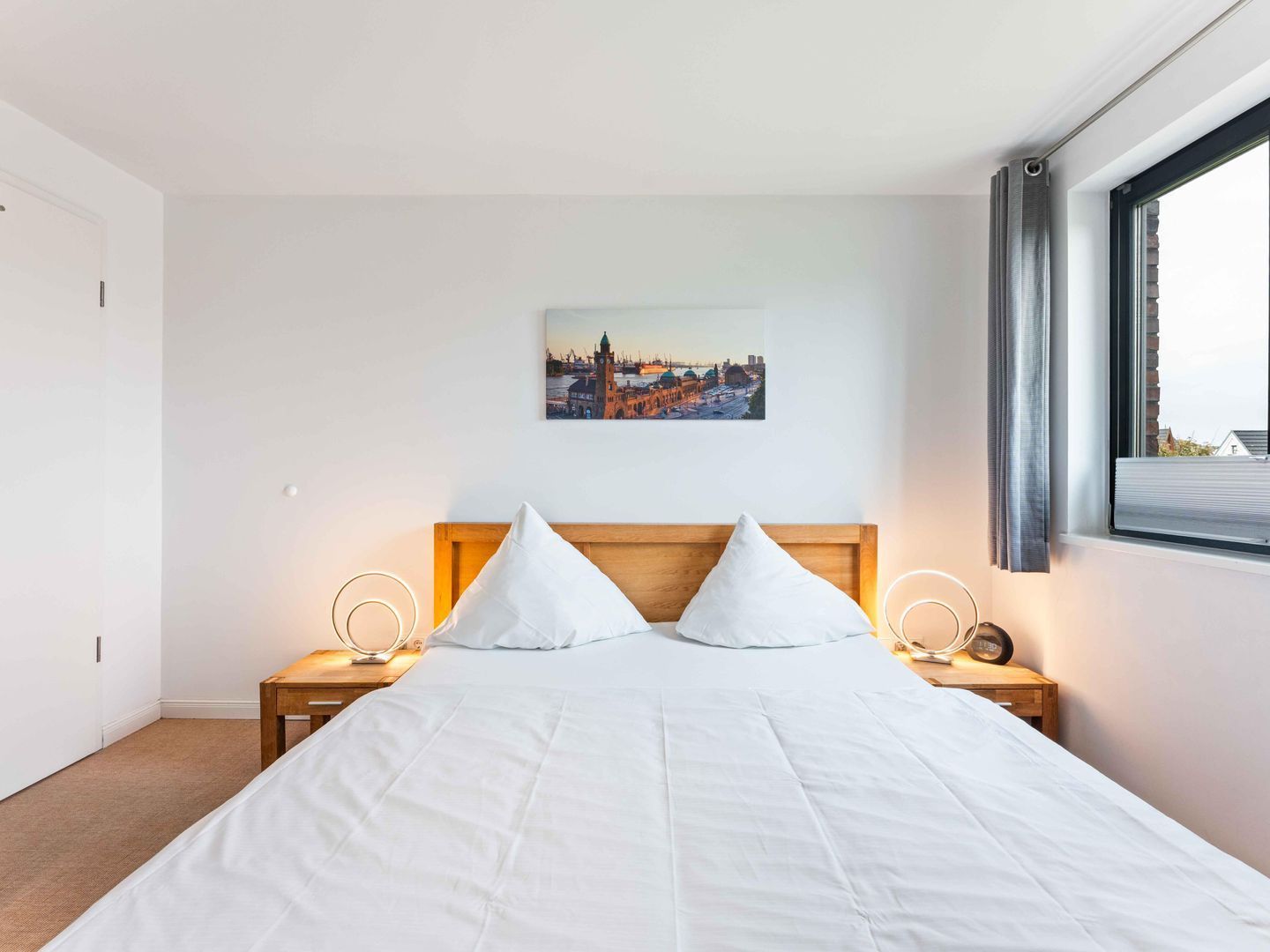 Une chambre d’hôtel avec un lit, deux tables de chevet, une commode, une fenêtre avec des rideaux et un tableau représentant un paysage urbain accroché au mur.