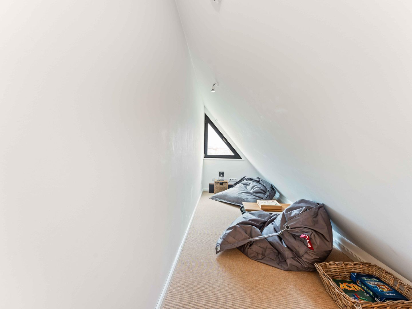 A narrow, sloped room with a high ceiling, a triangular window, and a small seating area with cushions and a basket of books.