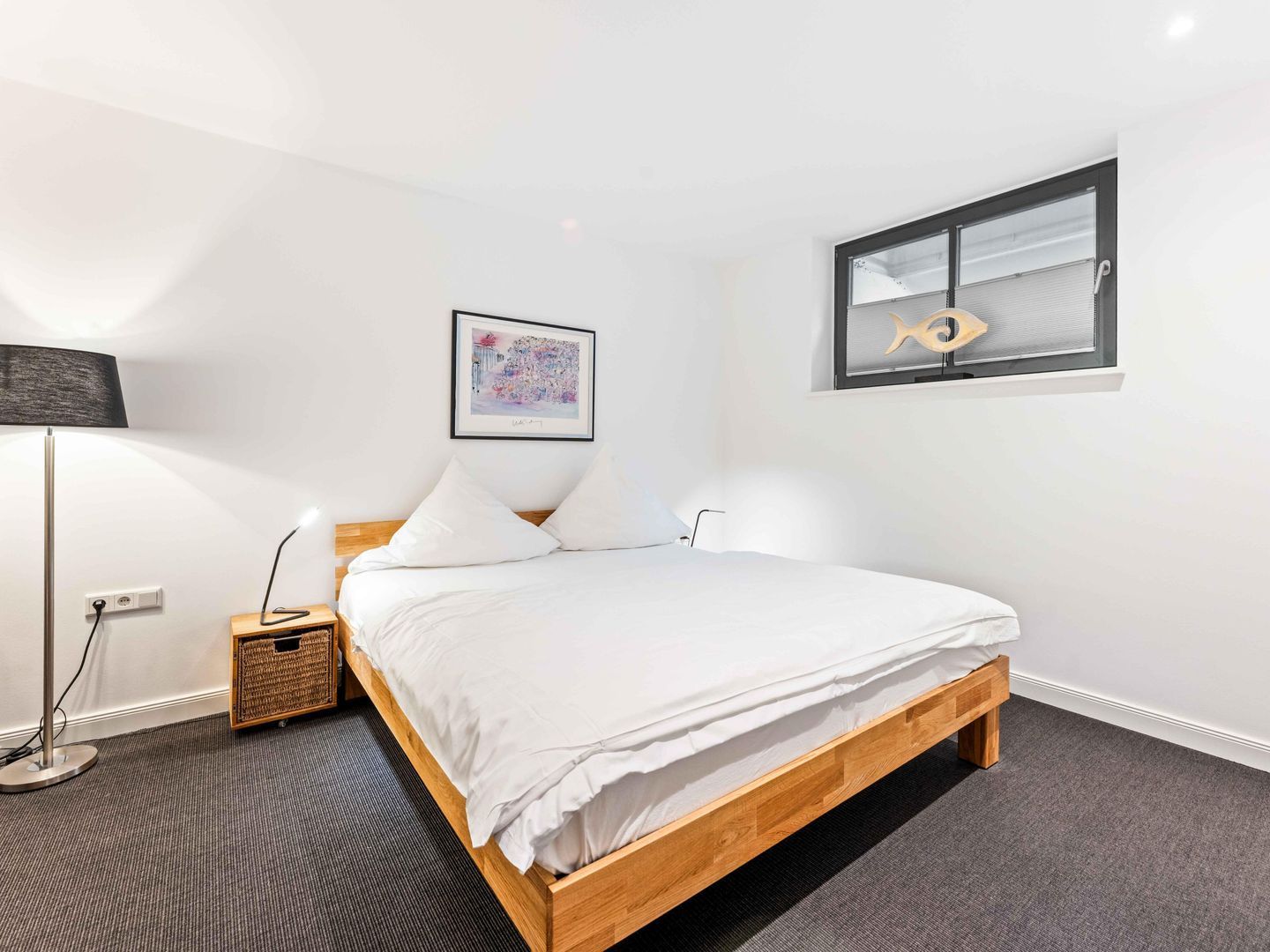 A bedroom with a wooden bed, white bedding, a floor lamp, a small bedside table, a framed picture, and a window with blinds.