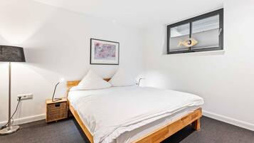 A bedroom with a wooden bed, white bedding, a floor lamp, a small bedside table, a framed picture, and a window with blinds.