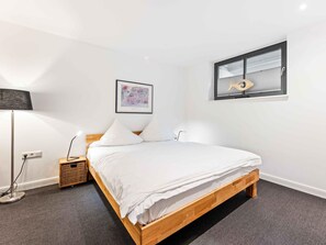 Un dormitorio con cama de madera, ropa de cama blanca, una lámpara de pie, una mesita de noche pequeña, un cuadro enmarcado y una ventana con persianas.