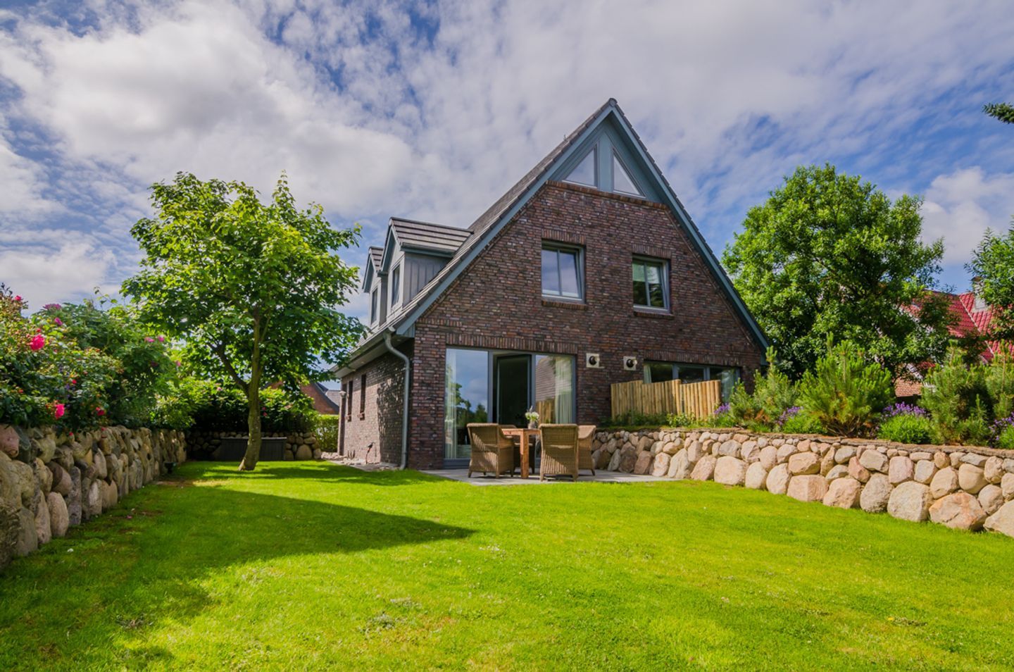 A two-story brick house with a sloped roof, a stone wall, and a well-maintained lawn.