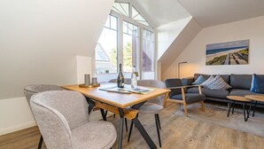 A modern living room with a wooden dining table, grey upholstered chairs, a grey sofa, and a painting of a beach scene. - Austernfischer App. 4 - 3-room apartment for up to 4 people with balcony. (Sylt)