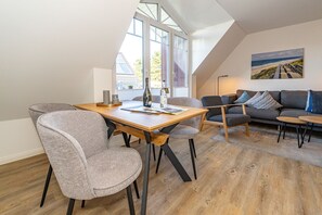A modern living room with a wooden dining table, grey upholstered chairs, a grey sofa, and a painting of a beach scene.