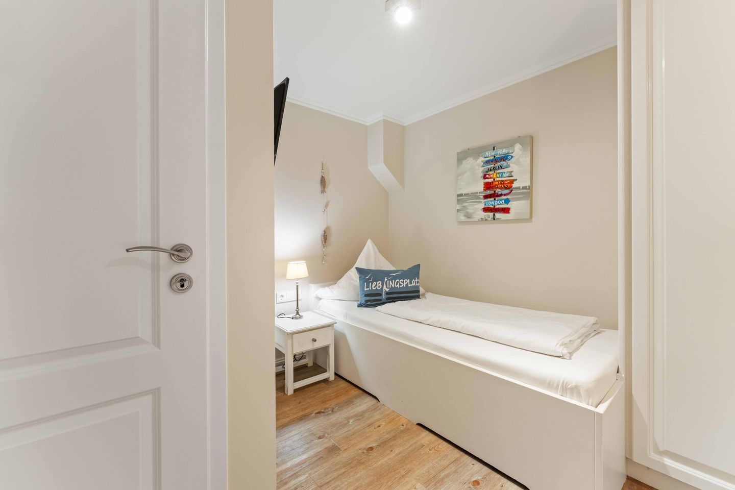 A small, neatly arranged bedroom with a single bed, a bedside table with a lamp, and a wall-mounted sign.