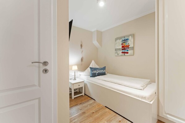 A small, neatly arranged bedroom with a single bed, a bedside table with a lamp, and a wall-mounted sign.