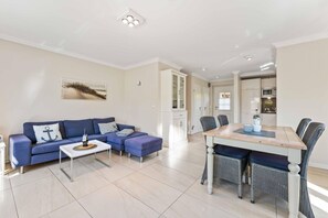 A modern living room with a blue sofa, a coffee table, and a dining area with a wooden table and wicker chairs.