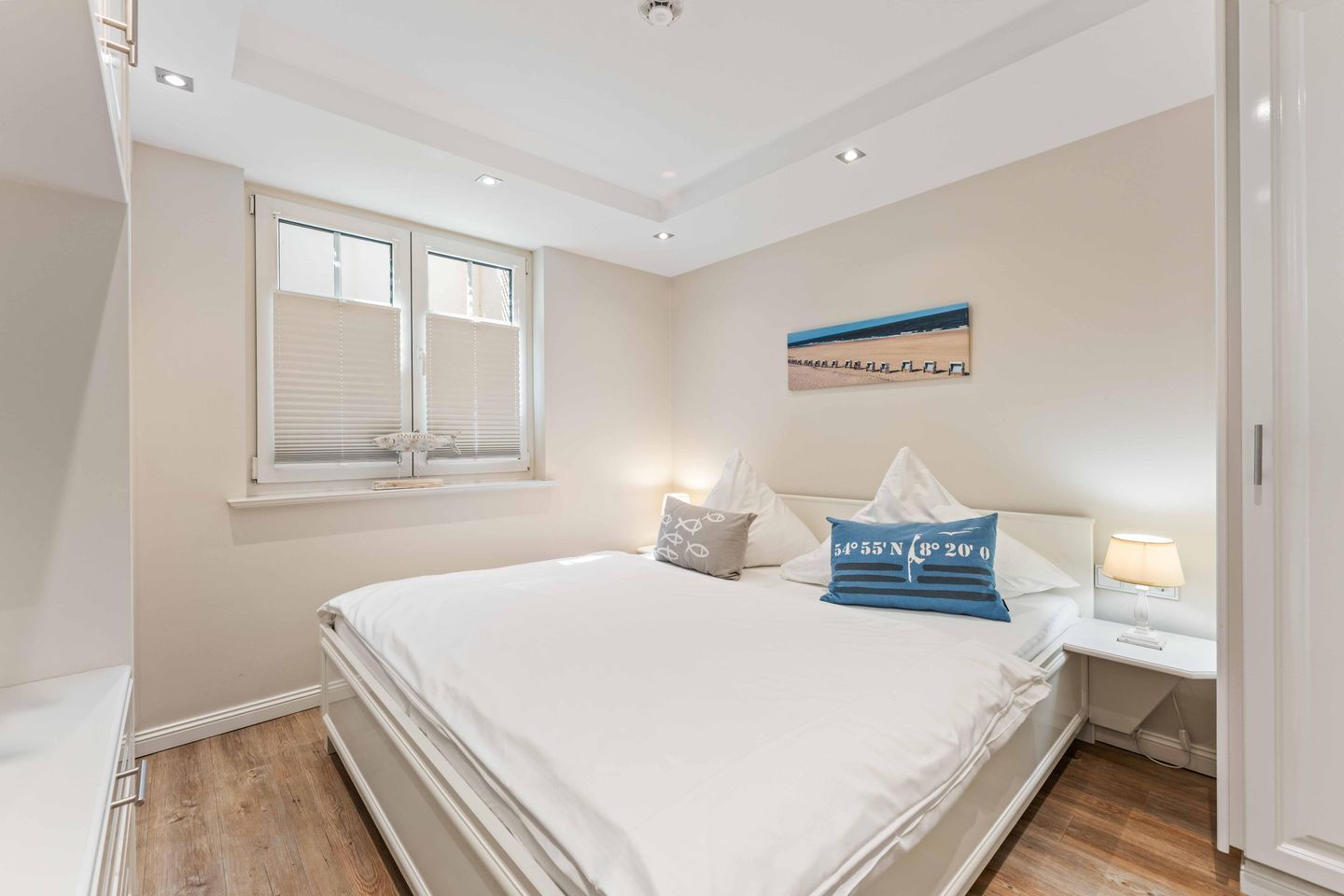 A bedroom with a large bed, a window with blinds, a nightstand with a lamp, and a wall-mounted picture.