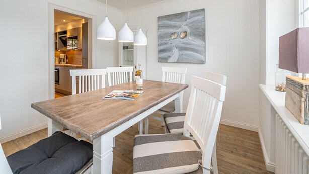 A dining area with a wooden table, white chairs, and pendant lights. A magazine is on the table, and a bottle of water is on the right. - Nordersuite - Just behind the dunes. 4-room vacation apartment in the north of (Sylt)