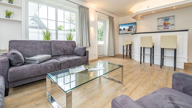 Un salon moderne avec une table basse en verre, un canapé gris et un coin cuisine doté de tabourets de bar.