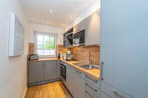 Eine kleine Küche mit grauen Schränken, einer Holzarbeitsplatte, einem eingebauten Backofen und einem Fenster mit Jalousien.