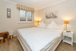A neatly made bed with white linens, a bedside table with a lamp, a small stool, a window with striped valance, and a wall-mounted painting of seashells. - Auszeit Sylt App. 2 - Hier ist der Name Programm, Nehmen Sie Sich Eine Auszeit auf Sylt (Sylt)