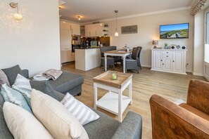 A cozy living room with a grey sofa, a wooden coffee table, and a brown armchair. A kitchen with white cabinets and a dining area with a table and chairs are visible in the background. - Auszeit Sylt App. 2 - Hier ist der Name Programm, Nehmen Sie Sich Eine Auszeit auf Sylt (Sylt)