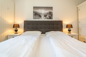 A double bed with white linens, two bedside lamps, a grey upholstered headboard, and a black and white landscape painting above the bed.