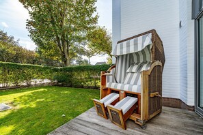A wooden deck chair with striped cushions and a woven canopy, positioned on a wooden deck in front of a white building.