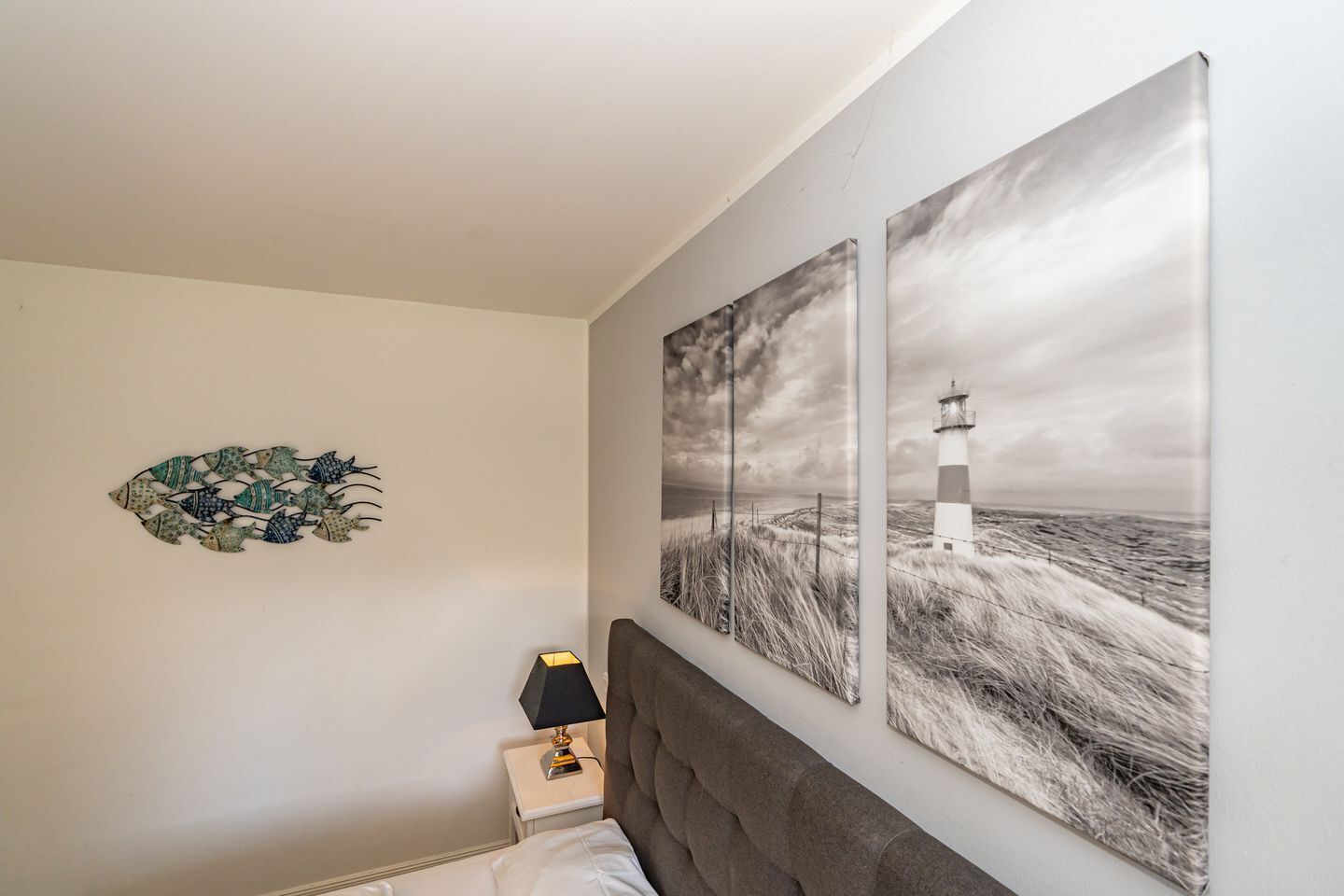 Ein Schlafzimmer mit einem Bett, einem Nachttisch, einer Lampe sowie einem Wandbild mit Fischmotiv und zwei Landschaftsbildern.