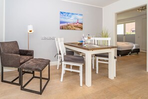 A modern, well-lit room with a wooden dining table, white chairs, a brown leather sofa, and a small bench. A painting of a lighthouse is on the wall, and a lamp is on the floor.