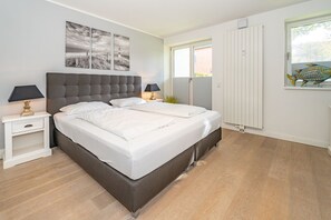 A modern bedroom with a large bed, two bedside tables with lamps, a window with blinds, and a wall-mounted artwork.
