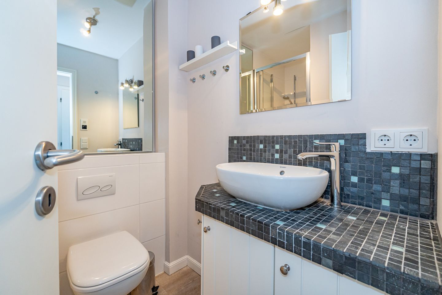 Et moderne badeværelse med et hvidt toilet, en stor vask med et spejl over og et flisebælte bag vasken.