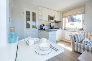 A compact kitchen with white cabinets, a microwave, and a coffee maker. There is a dining table with white plates and cups, and a striped sofa in the living area.