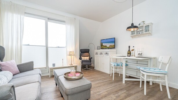 A modern living room with a grey sofa, a small table with a tray, a white dining table with chairs, and a TV mounted on the wall.
